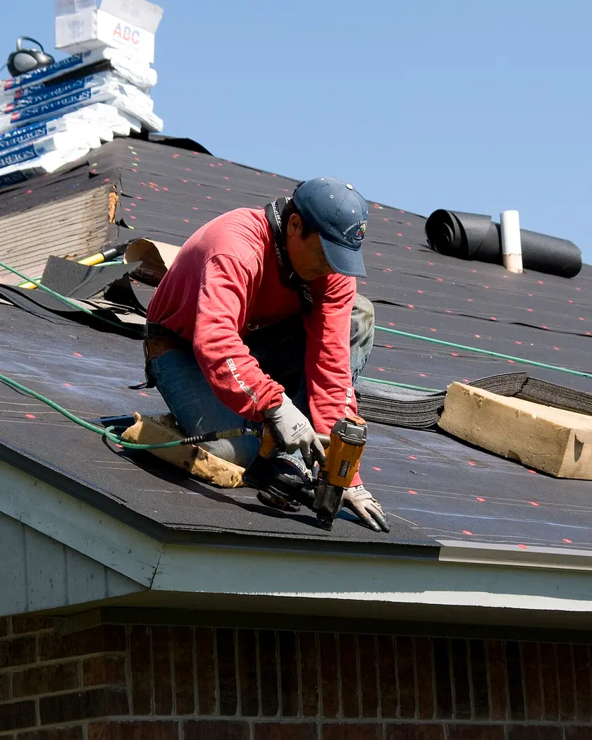 Roofer nailing underlayment on a house roof with stacks of shingles behind